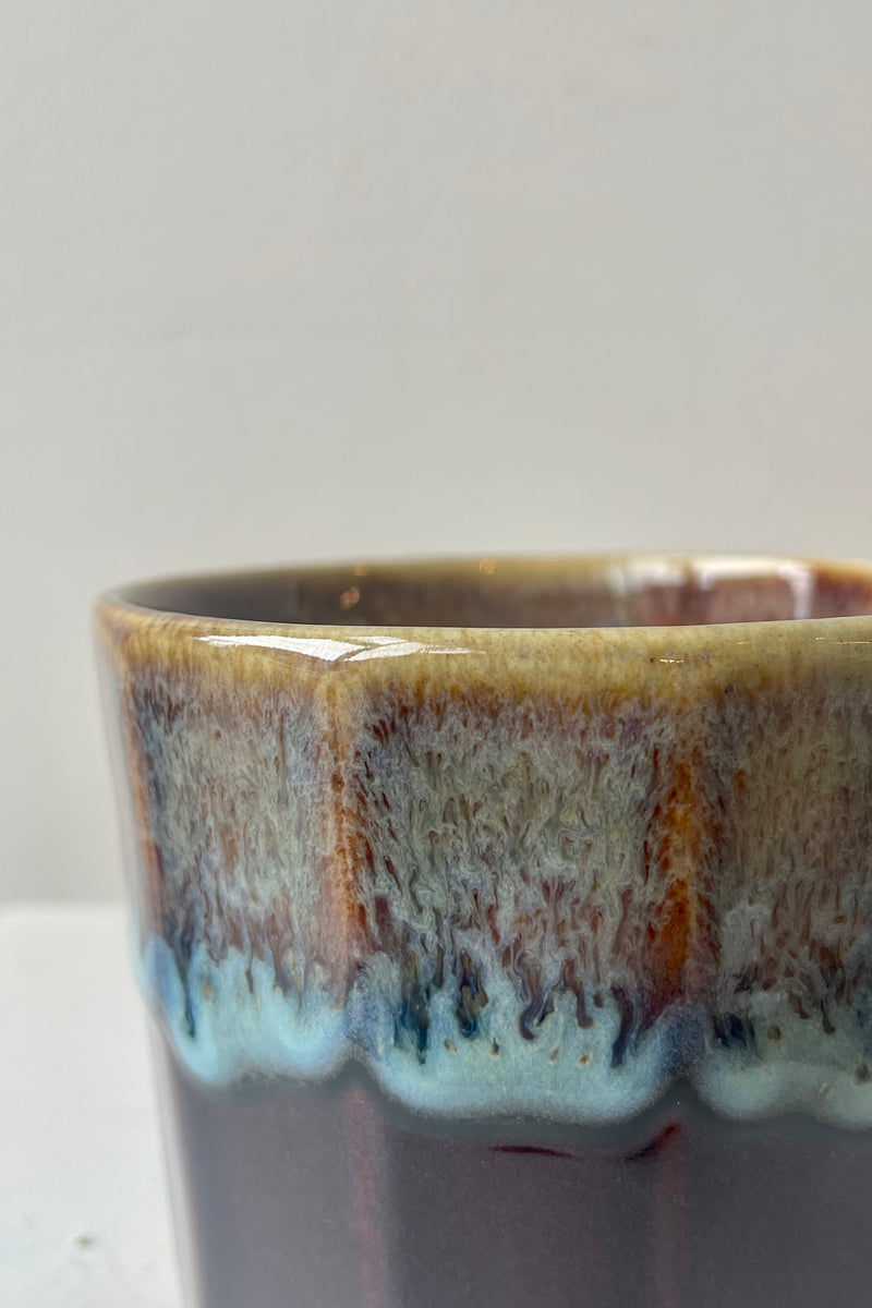 Close photo of amber, brown and blue glaze over black glazed mug against a white wall.