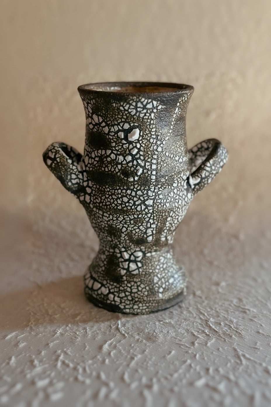 Grecian Crackle Vase in Dark Brown with a textured, crackled glaze pattern in black and white, featuring two small side handles, set against a neutral beige background. ©Olivia Snow Ceramics