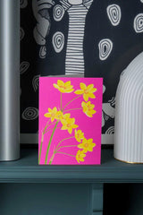 Daffodils greeting card by Stengun Drawings on a shelf with its bright yellow and fuchsia against a patterned white and black wall paper. 