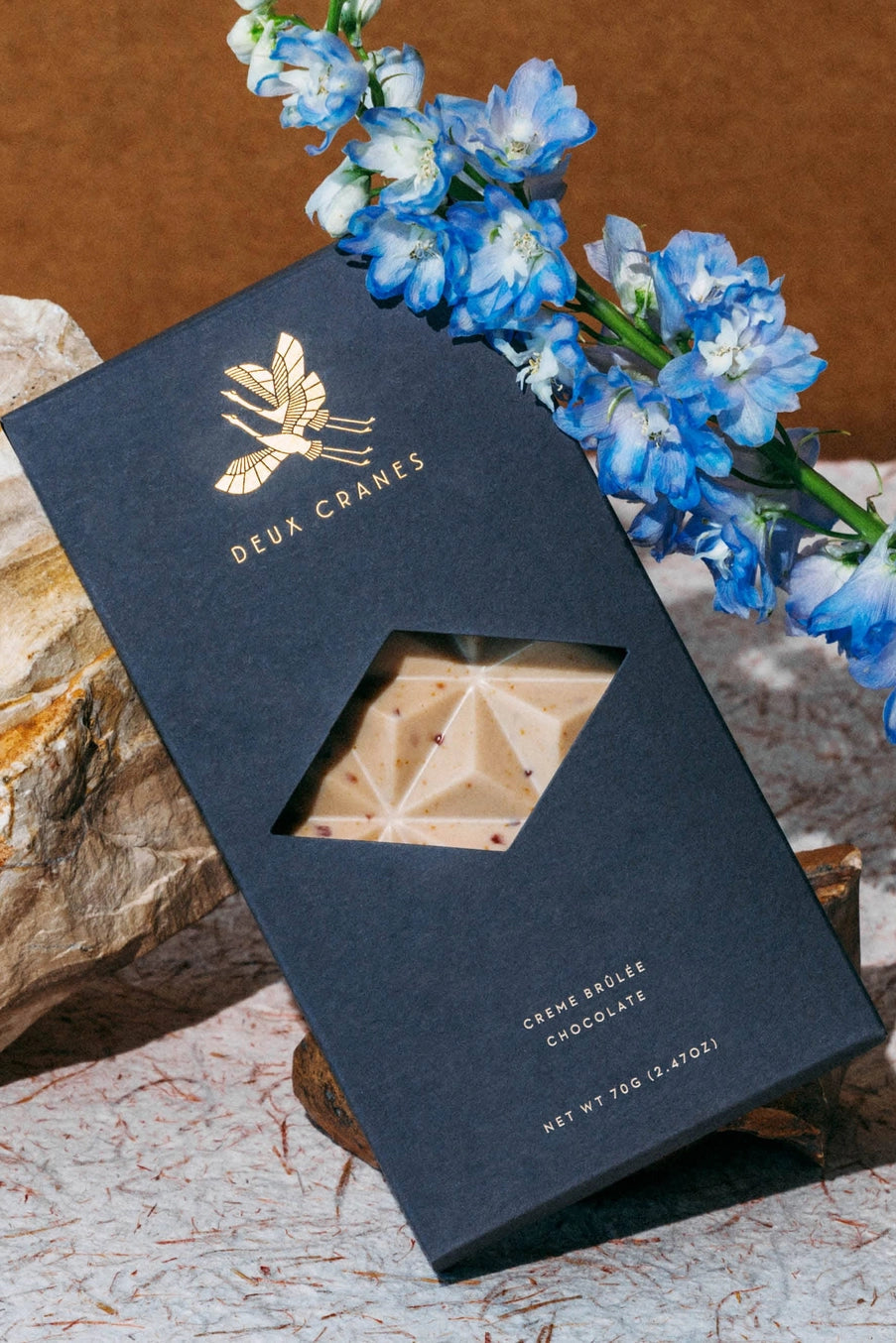 Creme Brûlée chocolate bar in a Deux Cranes packaging with blue flowers on a stone surface ©Deux Cranes