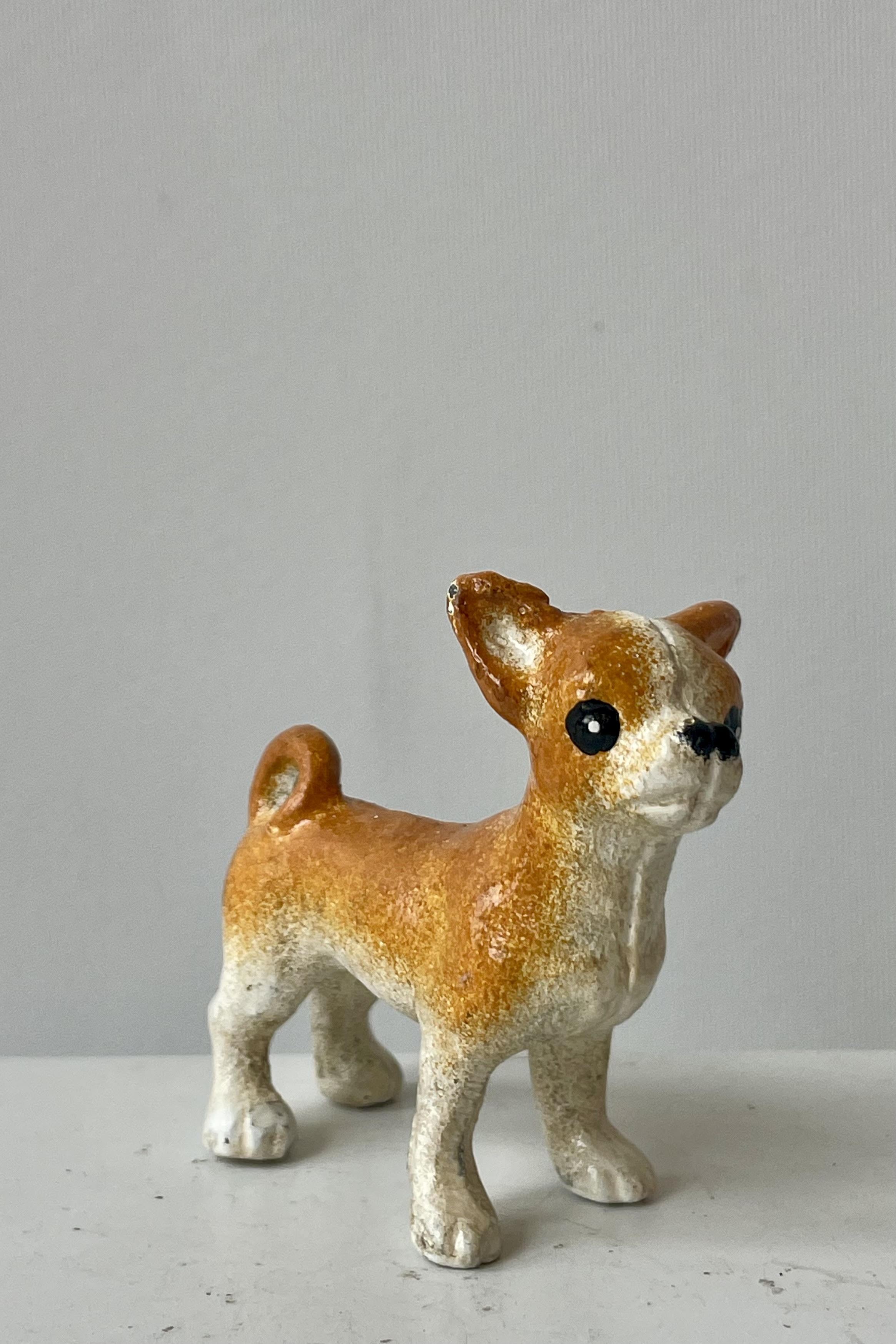 Photo of a white and tan Chihuahua figurine looking forward at an angle. It is shown on a white surface against a white wall.