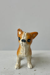 Photo of a white and tan Chihuahua figurine looking toward the viewer. It is shown on a white surface against a white wall.