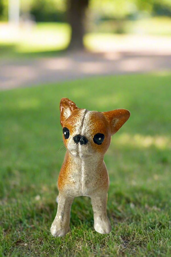 Photo of a white and tan Chihuahua figurine looking toward the viewer. It is shown in a park setting on grass.