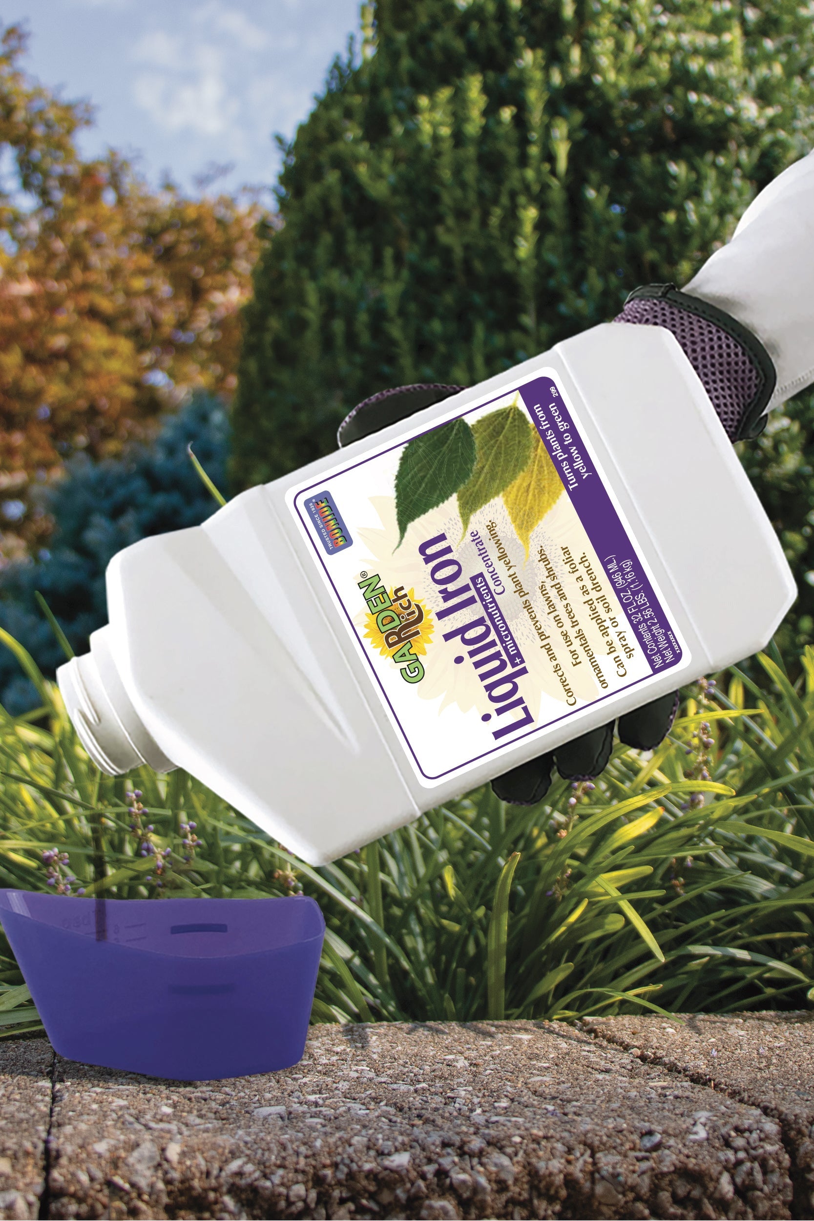 A hand wearing a white sleeve and purple glove holds a white bottle of Liquidon Plant Health liquid fertilizer and pours it into a purple measuring cup outdoors on a stone surface with green garden plants in the background. ©Bonide