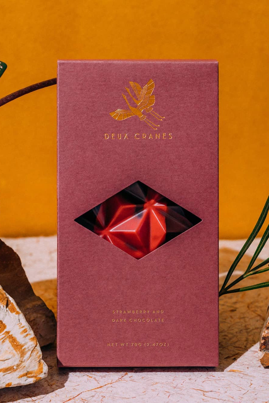 Pink chocolate box with a Dark Chocolate bar with strawberries on a decorative surface with plants and rocks. ©Deux Cranes