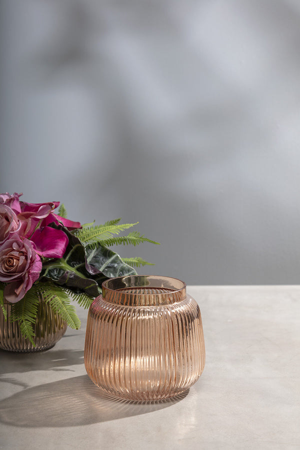 Ravish vase empty on an off-white table and gray background with another Ravish vase behind and off to the side with some flowers and ferns