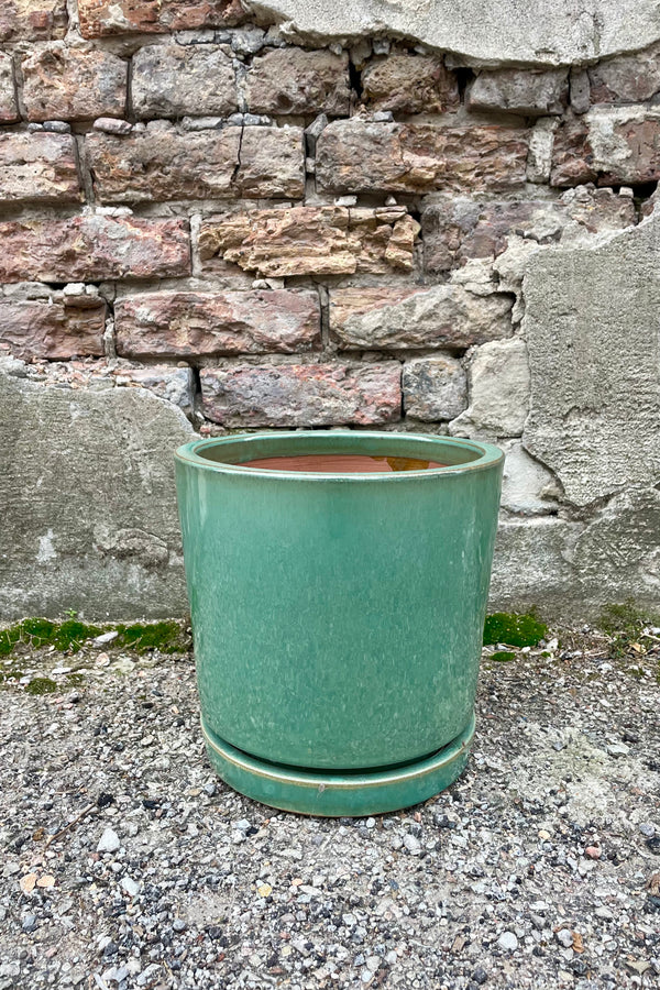 Photo of I/O2 sea green planter and saucer from the side. The semi-gloss glazed pot sits atop a fitted saucer. The photo is taken in front of a brick wall.