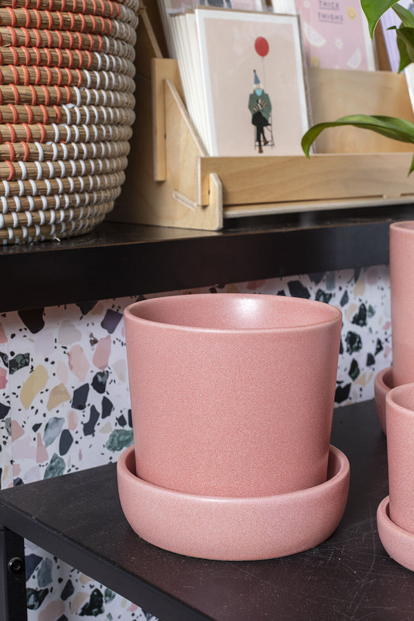 A pink Watson pot, empty, and sitting on a dark black table in front of a terrazzo wall and shelf with cards. 