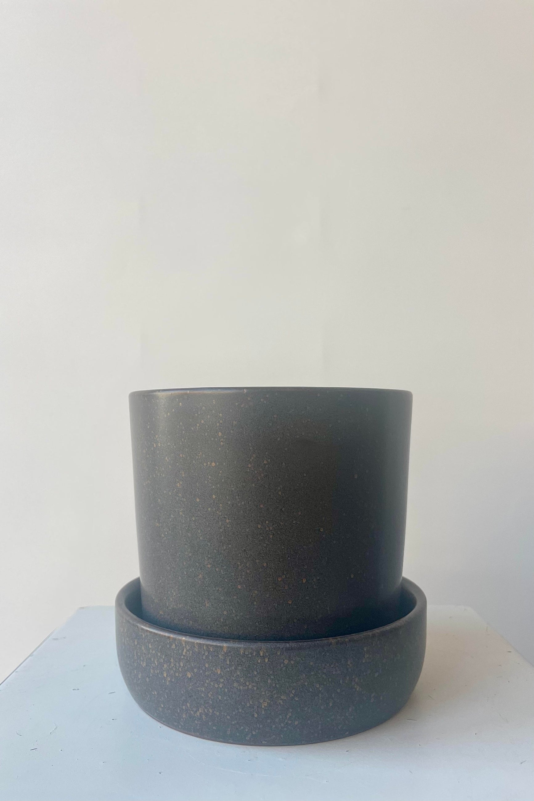 Watson Pot and Saucer with a soft black semi-gloss glaze with small flecks of clay showing through. It sits in a deep saucer and is shown in a white room on a white table ©Sprout Home