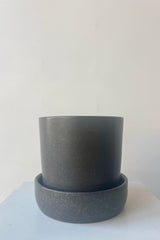 Photo of a Watson Pot and Saucer. The pot has a soft black semi-gloss glaze with small flecks of clay showing through. It since in a deep saucer and is shown in a white room on a white table.