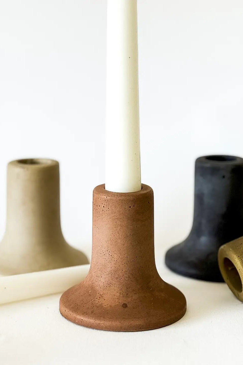 A set of matte ceramic candlestick holders in earthy tones, with a terracotta holder in the center holding a white candle, flanked by holders in beige, black, and olive green on a white background. ©Sunday Nomad