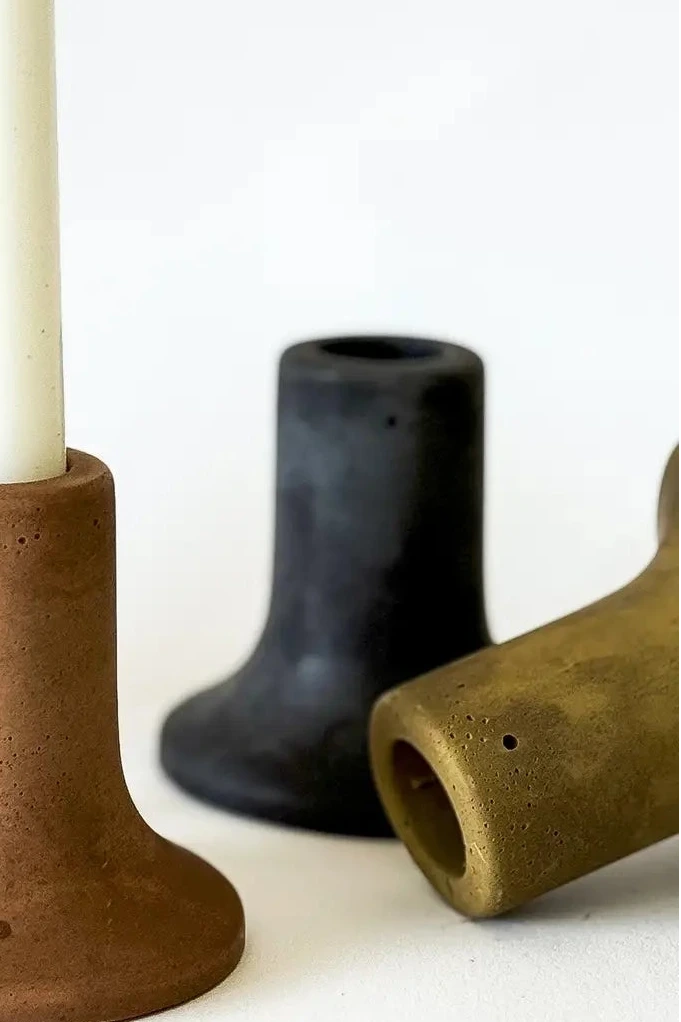 A set of matte ceramic candlestick holders in earthy tones, with a terracotta holder in the center holding a white candle, flanked by holders in beige, black, and olive green on a white background. ©Sunday Nomad