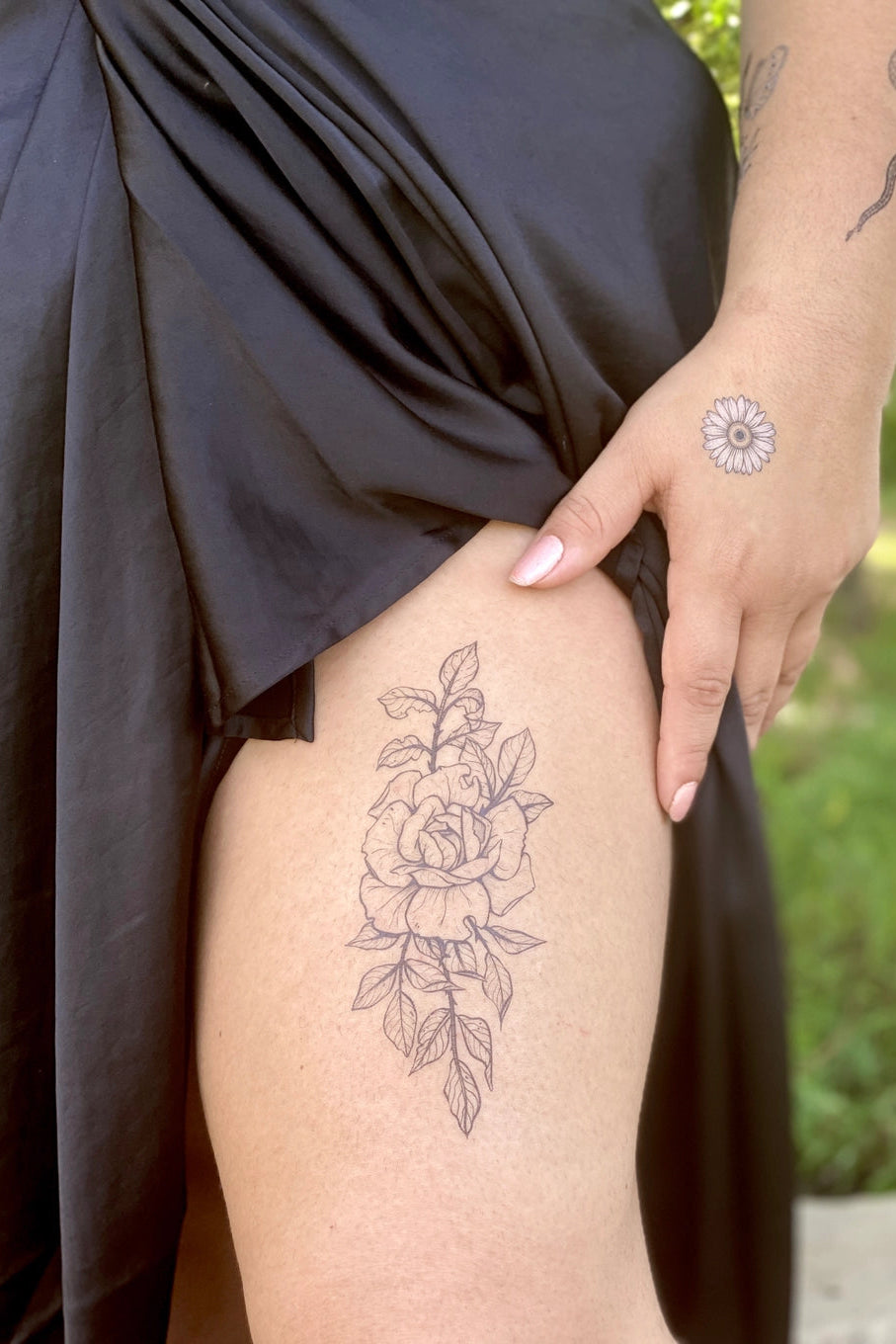 Person with Rose design floral tattoos on leg and hand, wearing a dark dress outdoors. ©Nature Tats