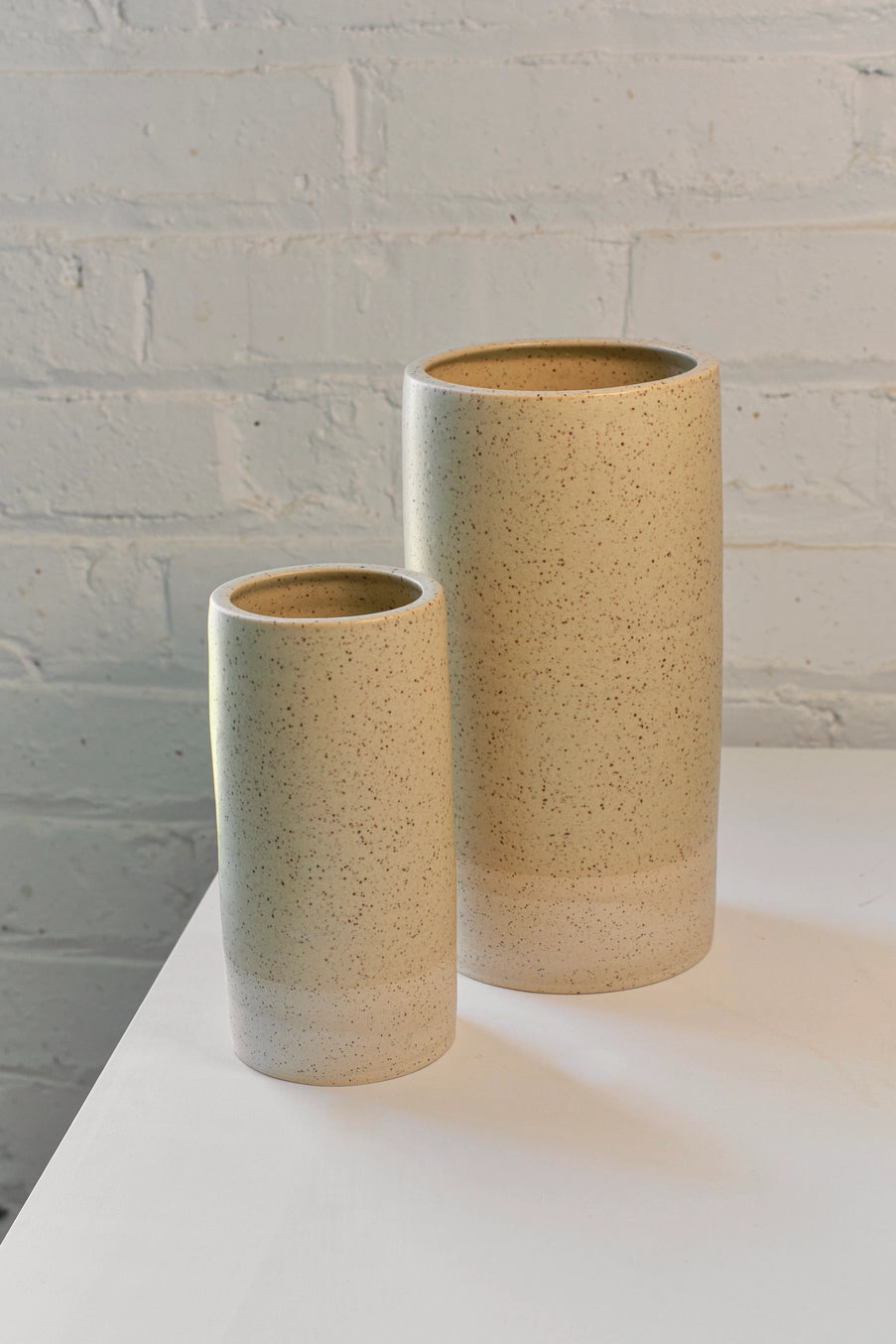 Two ceramic Butter Specked Jacqueline vases with a textured finish on a white surface against a light gray brick wall. ©Little Fire Ceramics