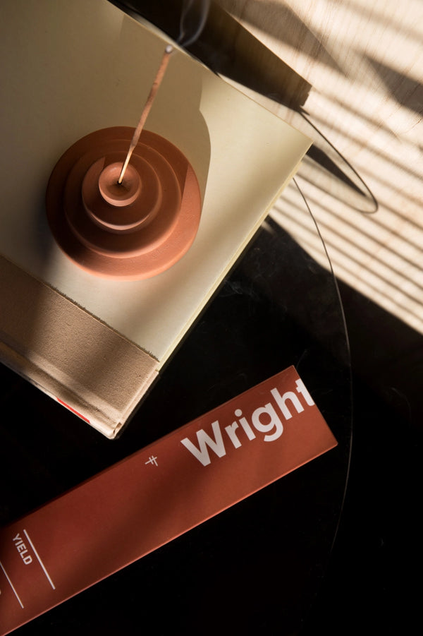 Wright Stick Incense by Yield  on a table in a dimly dramatic light room and a burning stick next to it on a book. 