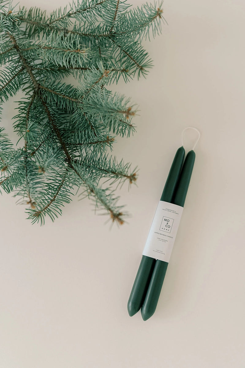 Mo & Co beeswax taper candles in Forest Green with an evergreen branch laying near.