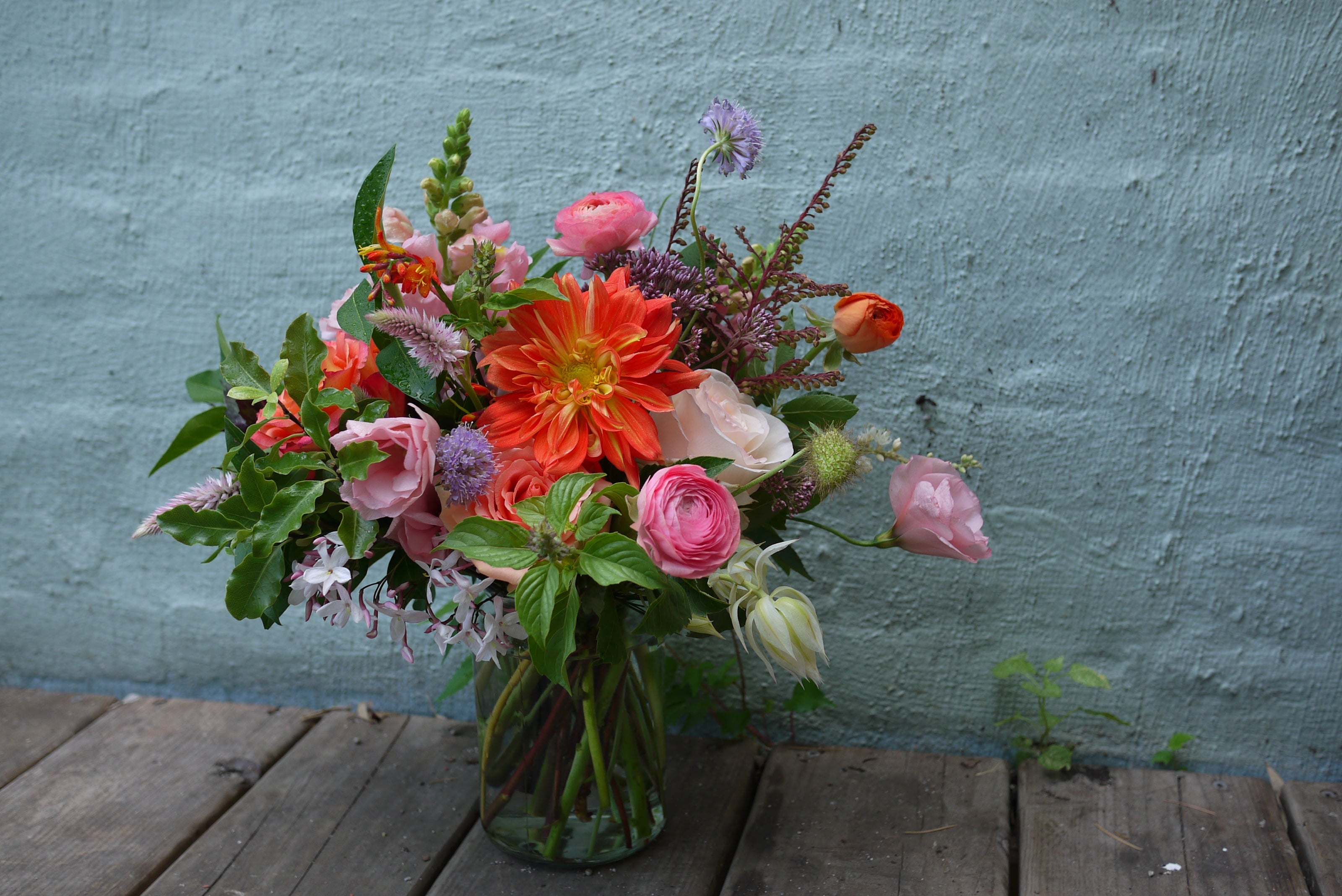 Bright bouquet of flowers including dahlia, ranunculus and celosia in a clear vase on a wooden table in front of a blue wall. 