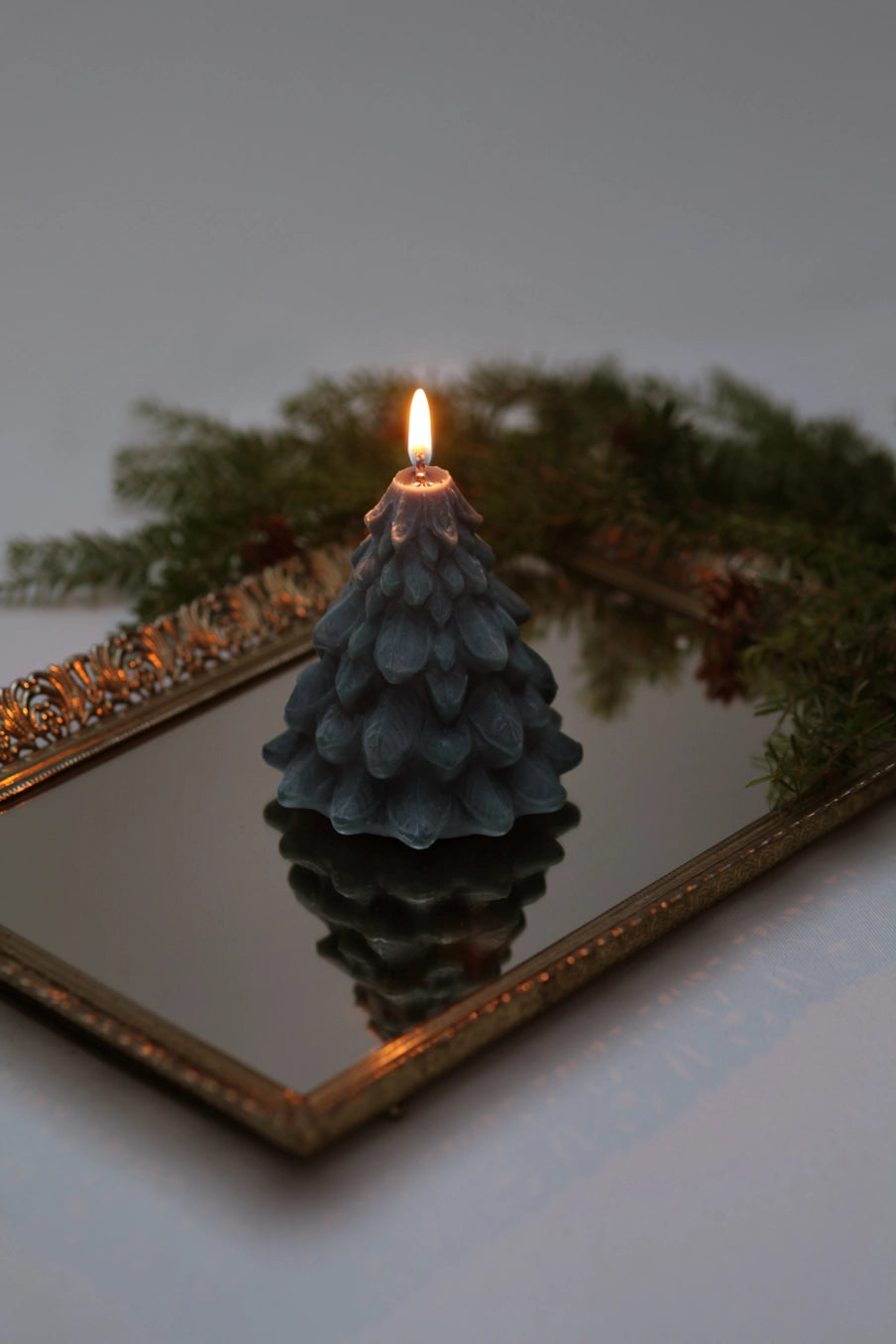 A tree-shaped teal teal candle with a lit flame sits on a decorative gold-framed mirror, surrounded by green pine garland. ©Mo&Co.