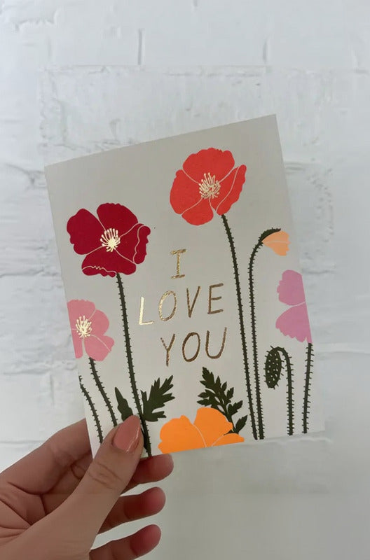 Card with red flowers and 'I LOVE YOU' text held against a white brick wall. ©Hartland Cards