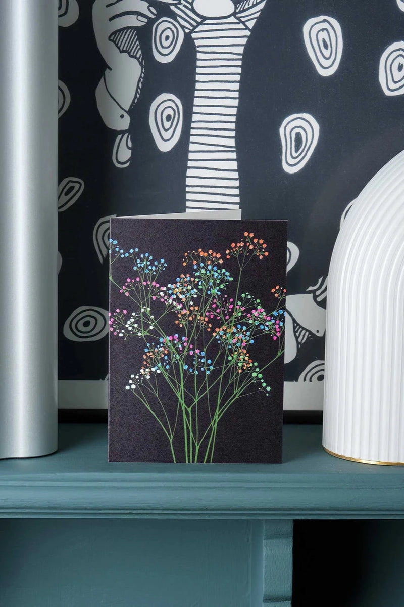 Rainbow Gypsophila greeting card by Stengun Drawings in a lifestyle shot on a ledge and black and white wallpaper behind the card. 