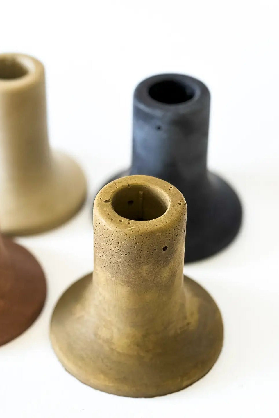 Four small cone-shaped cement candle holders with flared bases in terracotta, beige, charcoal, and ochre tones, arranged on a plain white background. ©Sunday Nomad