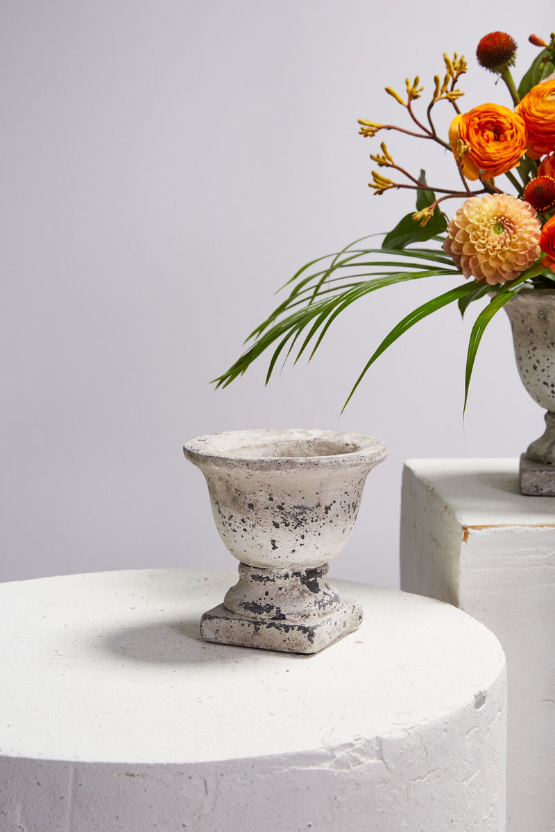 The Sanctuary Urn sitting on a white concrete table in front of a partially viewed floral arrangement in orange. 