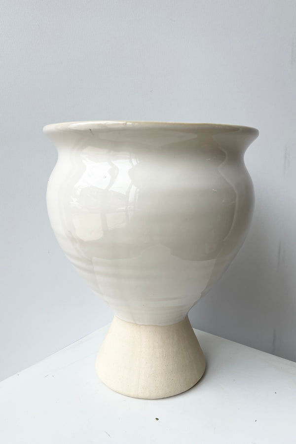 The Magnolia Vase large against a white wall viewed at eye level from the side showing its urn like shape and white glaze. 