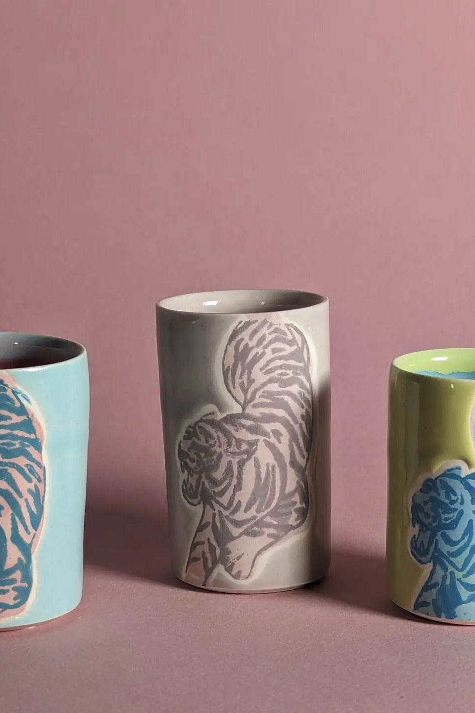 Three handmade ceramic tumblers with raised animal designs, displayed against a solid dusty pink background. The left tumbler is light blue with a pink tiger design, the middle is light gray with a gray-brown tiger design, and the right is lime green with a blue tiger design. ©City In Bloom Ceramics