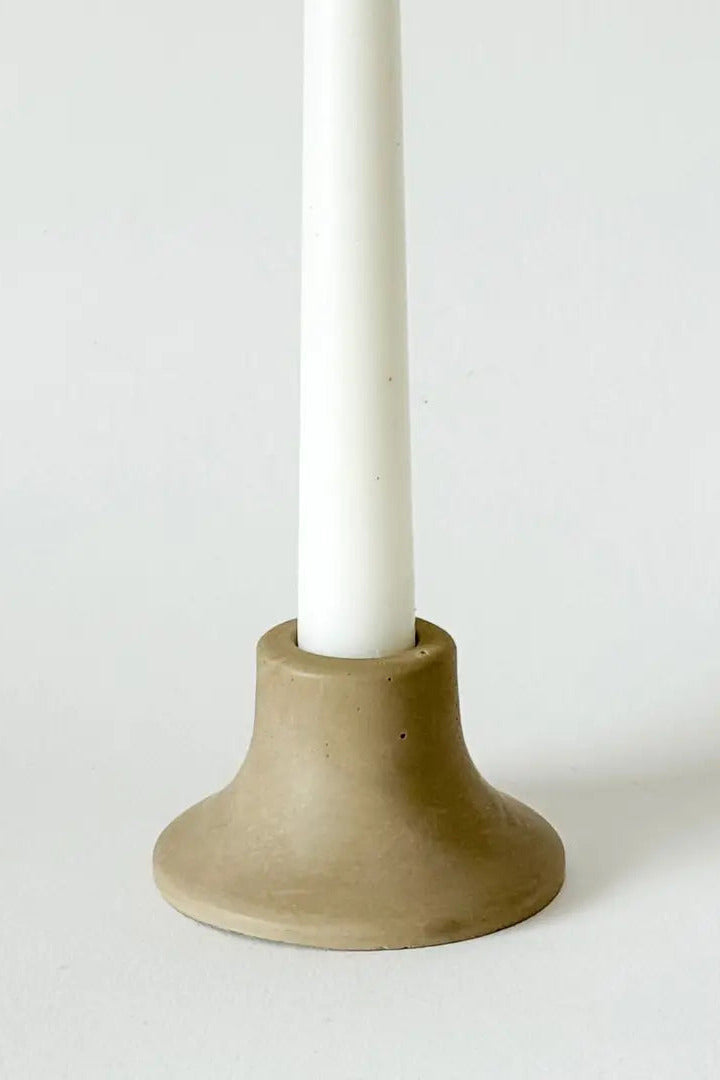 A minimalist ceramic candle holder with a wide, flared base in a warm taupe-beige color, holding a white taper candle, set against a clean white background. ©Sunday Nomad 