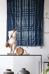 a tie dyed indigo mud cloth hanging on a white wall in front of a shelf with a sculpture n it with metal and leather objects.