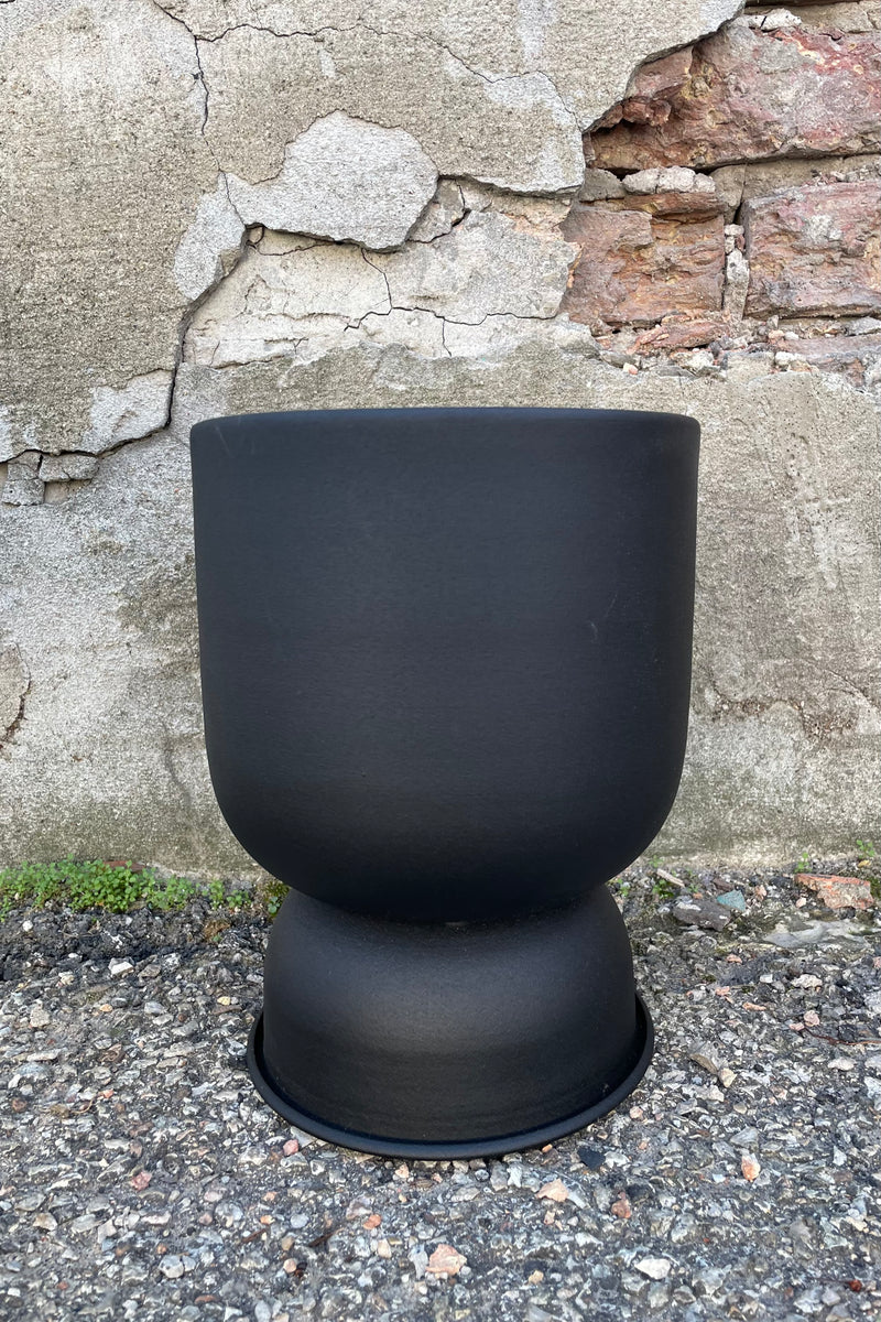 The 7.5" x 10" black metal compote pot viewed from the side eye level against a concrete wall. 