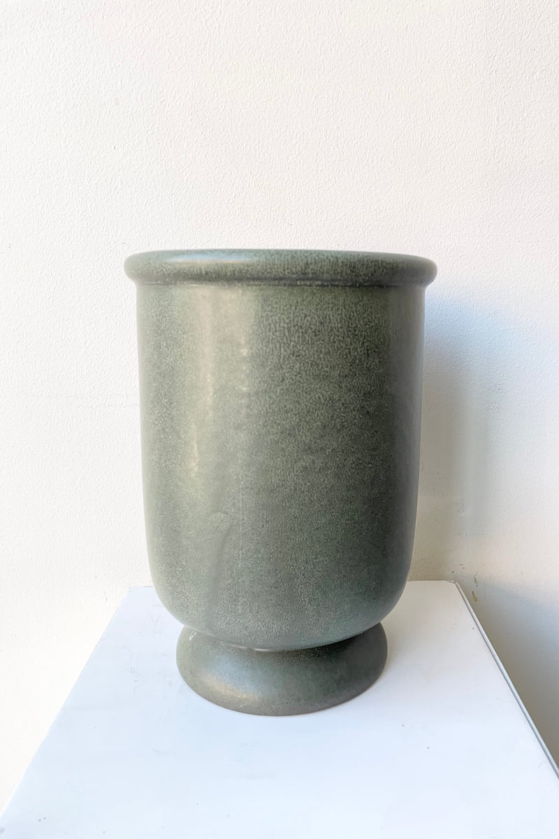 The dark green stoneware urn vase shown from the side against a white wall. 