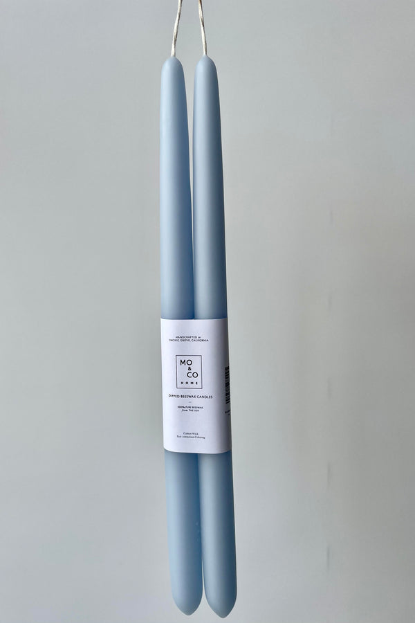 Photo of a pair of Mo & Co bee's wax taper candles. The color of the candles is Bluebell and they have a white wrapper with a black logo across the middle of the pair. They are photographed against a white wall.