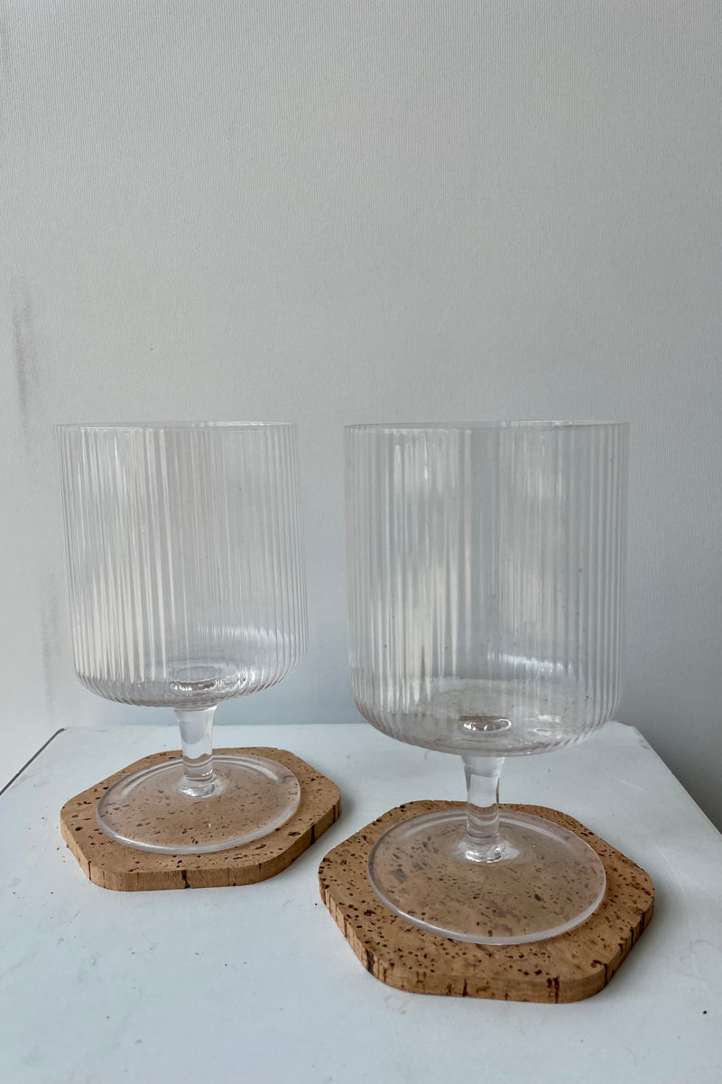 six-sided cork coasters are photographed on a white surface against a white wall. On each coaster is a clear glass stemmed wine glass.