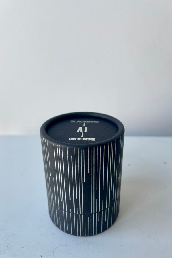 A cylinder of Blackbird incense sits on a white table in a white room. The incense is called Ai. Packaging features a black background with a silver detail of vertical parallel lines of varying lengths.