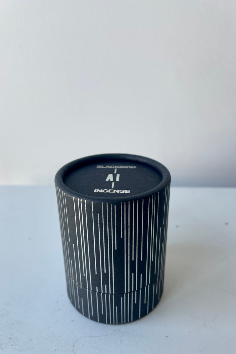 A cylinder of Blackbird incense sits on a white table in a white room. The incense is called Ai. Packaging features a black background with a silver detail of vertical parallel lines of varying lengths.