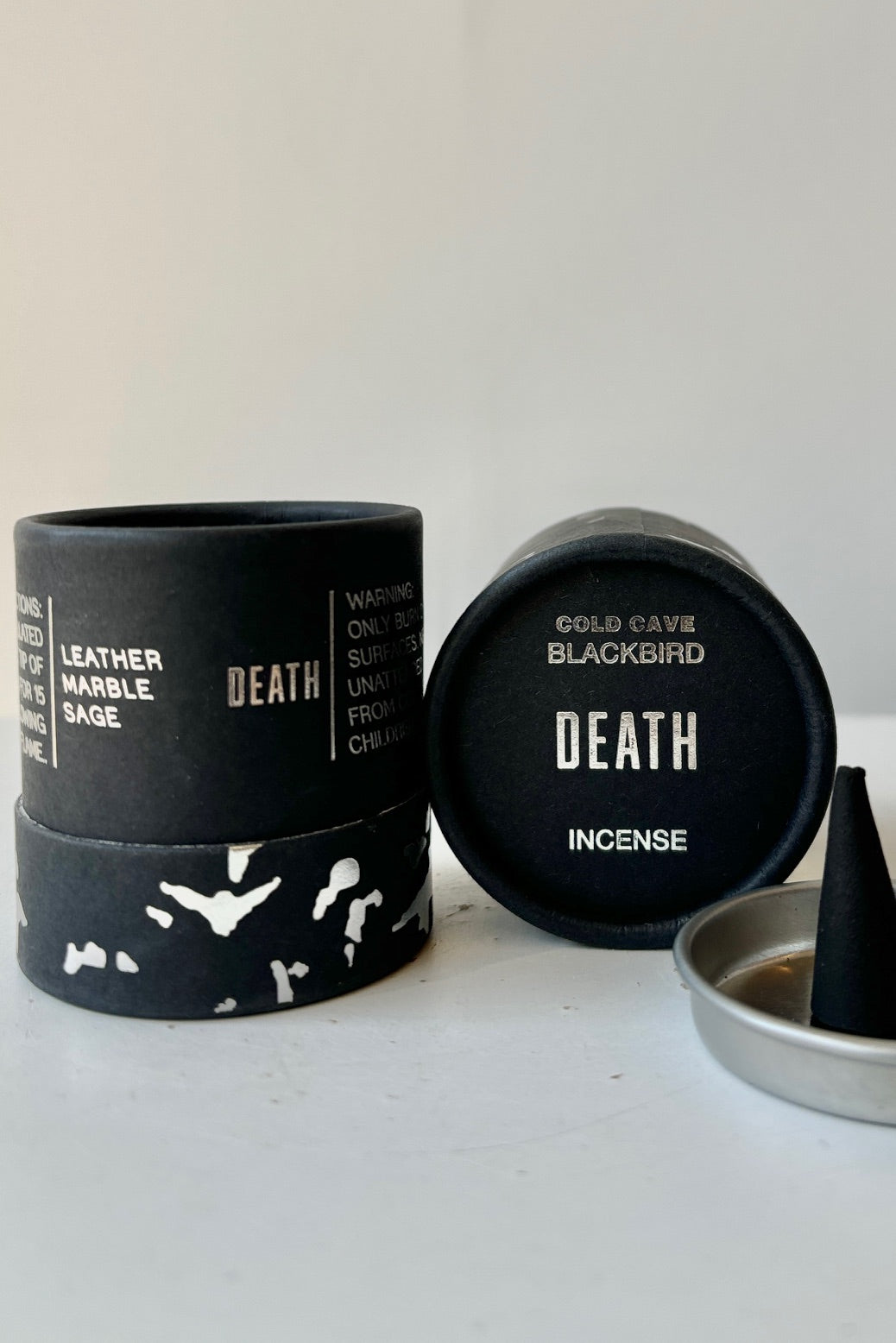 A black cardboard cylinder featuring silver patterning and type with a circular silver tray and one cone incense on a white table with white background. This scent is called Death and the fragrance contains notes of leather, marble and sage. ©Sprout Home