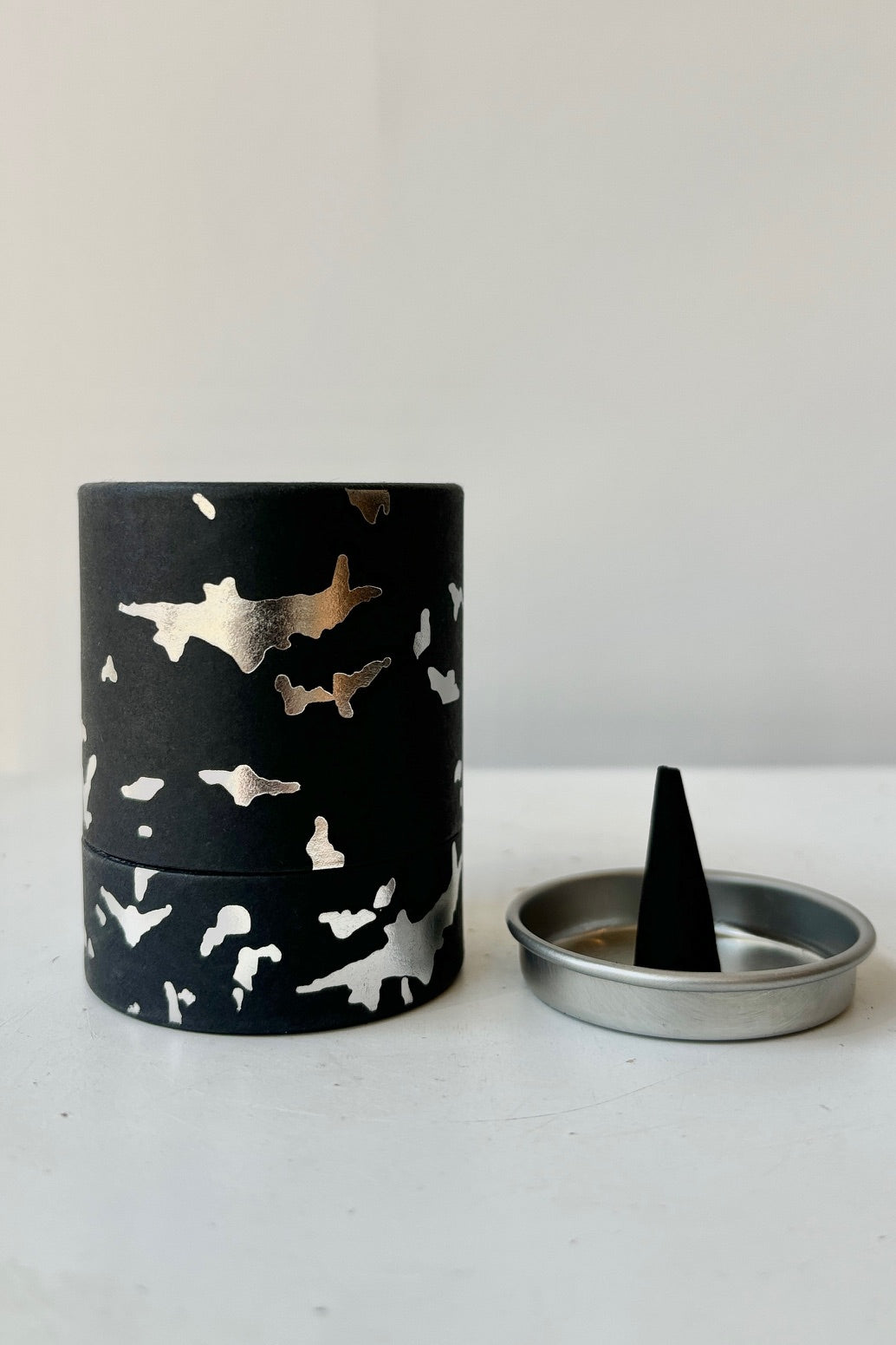 A black cardboard cylinder featuring silver patterning and type with a circular silver tray and one cone incense on a white table with white background. This scent is called Death and the fragrance contains notes of leather, marble and sage. ©Sprout Home