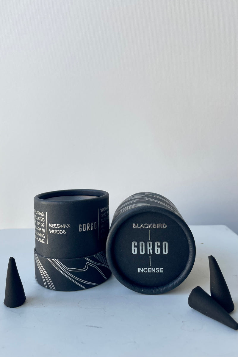 A black cylinder of Blackbird incense sits on a white surface in a white room. The package features undulating silver parallel lines. The incense is labelled Gorgo. To the left and right are pieces of Incense cones.