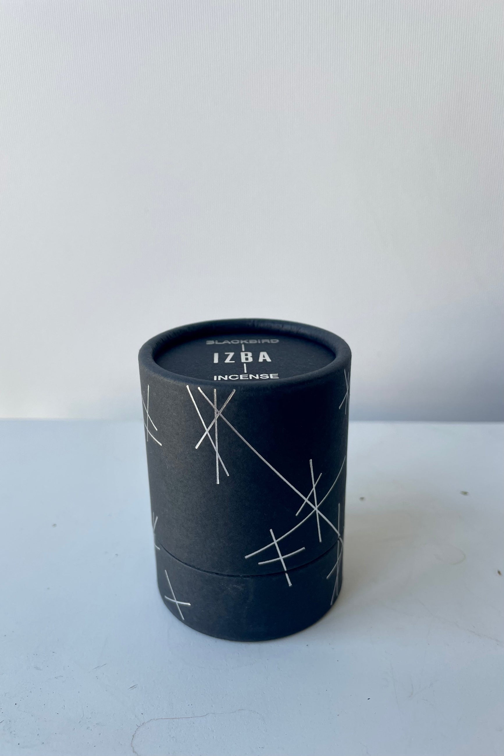 A black cylinder of Blackbird incense sits on a white surface in a white room. The package features a pattern of intersecting silver lines. The incense is called Izba. ©Sprout Home