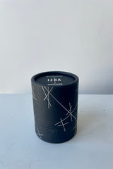 A black cylinder of Blackbird incense sits on a white surface in a white room. The package features a pattern of intersecting silver lines. The incense is called Izba.