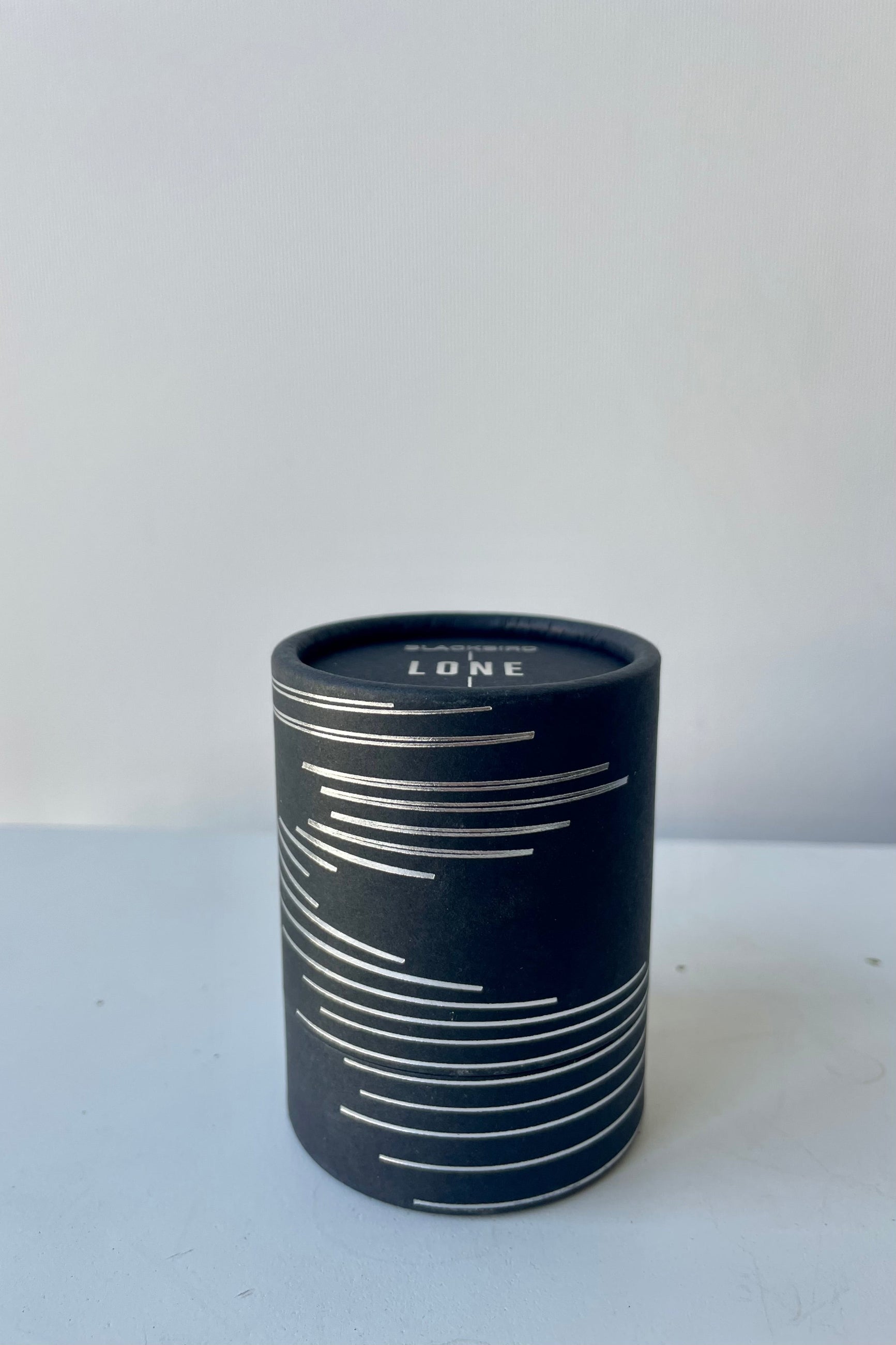 A black cylinder of Blackbird incense sits on a white surface in a white room. This incense is called Lone and the package features a silver lined design.  ©Sprout Home