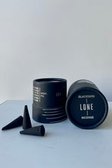 A black cylinder of Blackbird incense sits on a white surface in a white room. This incense is called Lone and the package features a silver lined design. To the left sits a scattering of black incense cones.