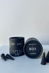 A black cylinder of Blackbird incense sits on a white table in a white room. The incense is called Muru. The packaging features a black background with silver lines and circles.  To the left and right and black incense cones.