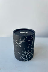 A black cylinder of Blackbird incense sits on a white table in a white room. The incense is called Muru. The packaging features a black background with silver lines and circles. 
