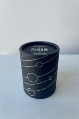 A black cylinder containing Blackbird Incense sits on a white table in a white room. The incense is called Ploom. Packaging features a black background with intersecting silver lines and circles.