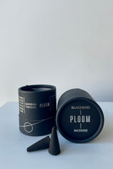 A black cylinder containing Blackbird Incense sits on a white table in a white room. The incense is called Ploom. Packaging features a black background with intersecting silver lines and circles. Ahead of it sits a pair of black incense cones.