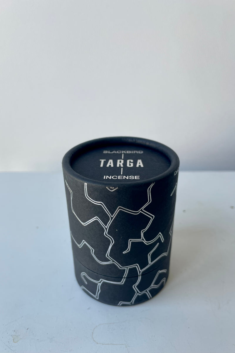 A black cylinder of Blackbird incense sits on a white table in a white room. The incense is called Targa. The packaging features parallel jagged silver lines.