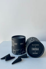 A black cylinder of Blackbird incense sits on a white table in a white room. The incense is called Targa. The packaging features parallel jagged silver lines. To the front and left sits a scattering of black incense cones.