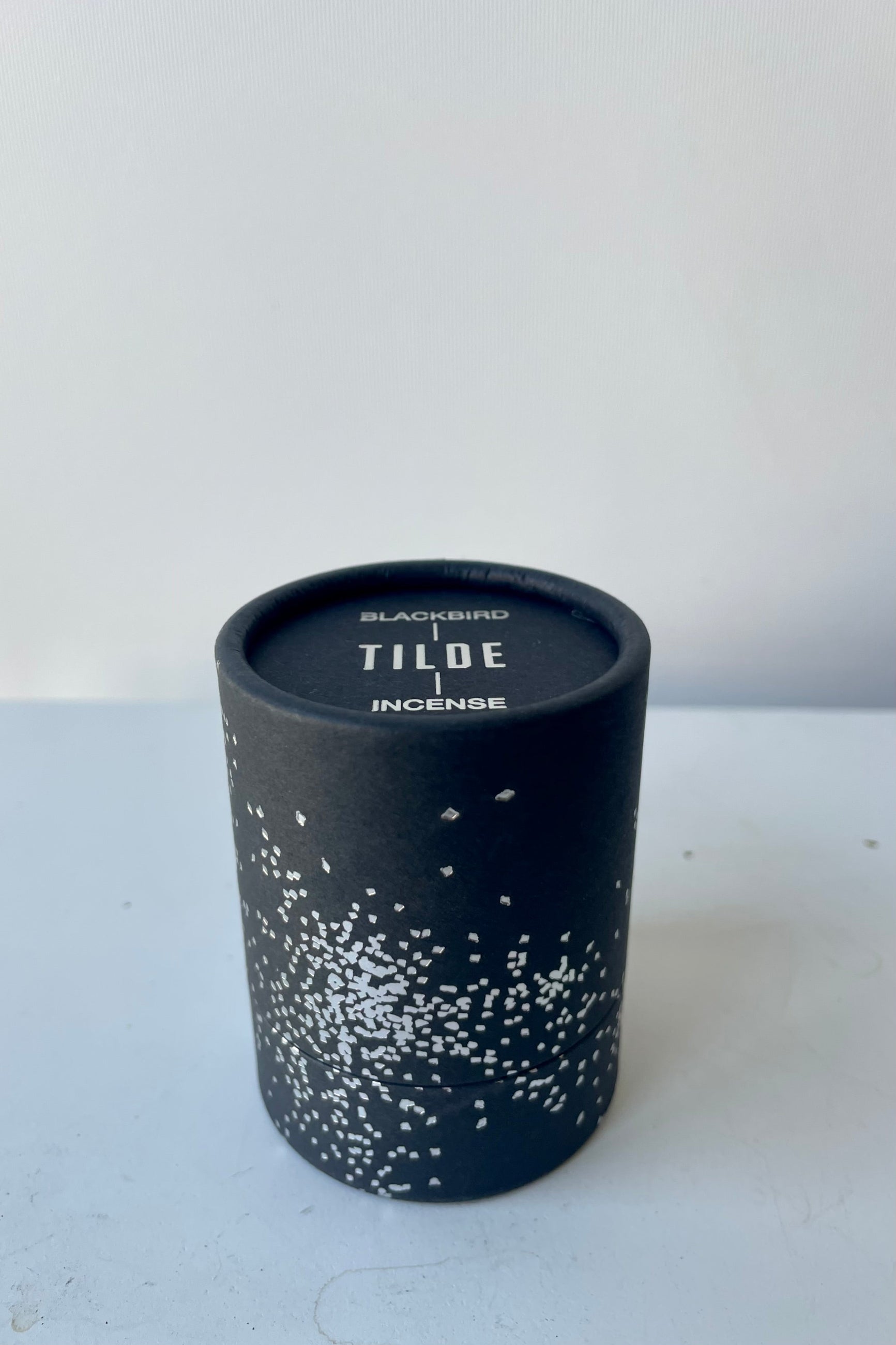 A cylinder of Blackbird incense sits on a white table in a white room. The incense is called Tilde. The packaging features a black background with silver detail of clusters of small irregular geometric shapes. ©Sprout Home